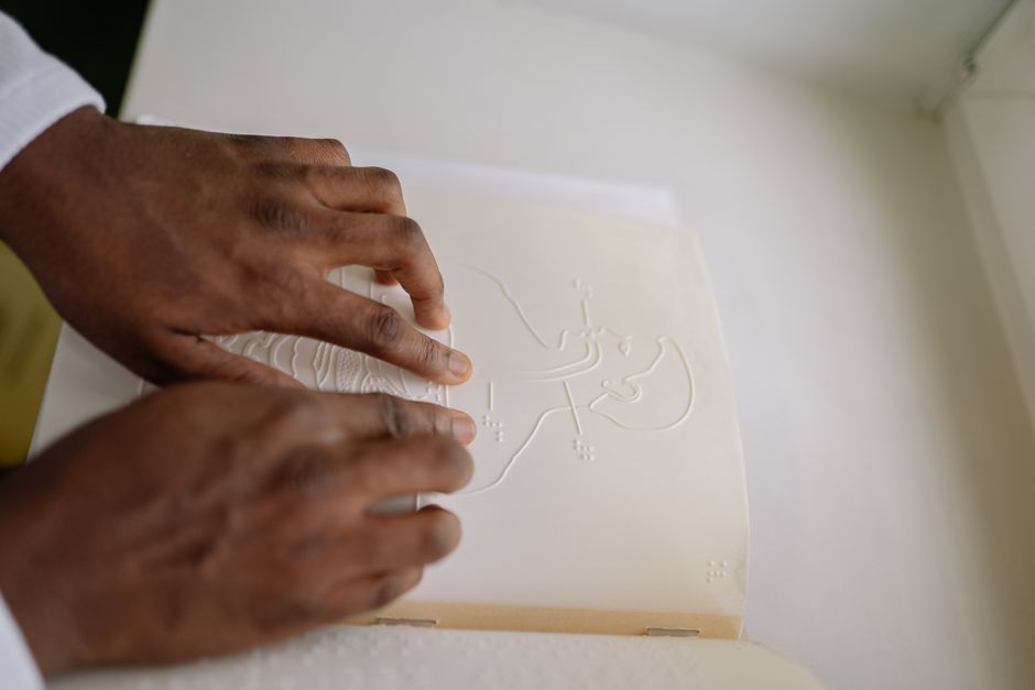 Acessibilidade Brasil — Close-up of hands reading a tactile, braille-embossed educational book indoors.
