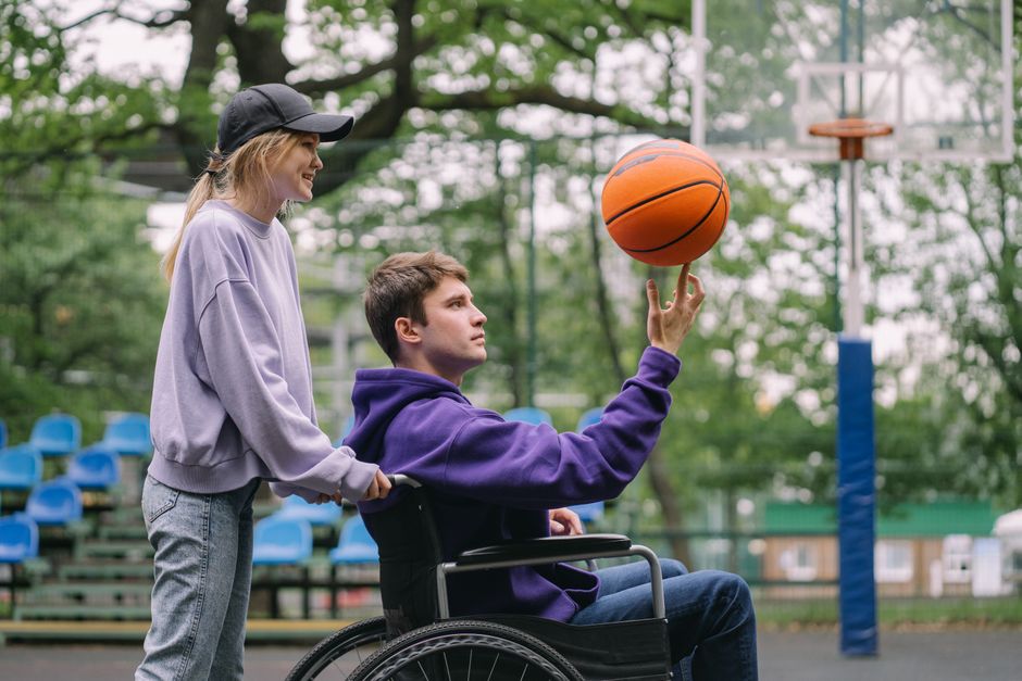 Acessibilidade Brasil — A young man in a wheelchair spins a basketball outdoors with a supportive woman by his side.