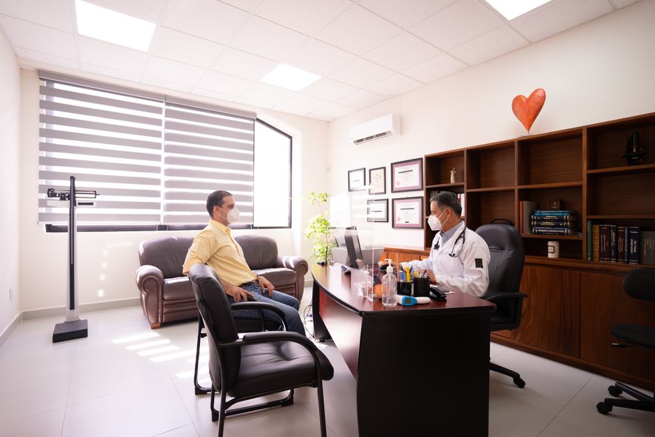 Acessibilidade Brasil — A patient consults with a masked doctor in a well-lit, modern office. Safety measures in place.